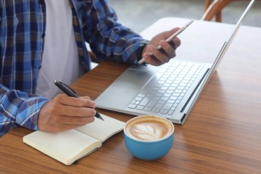 Erkek el yazısını kalemle deftere kapat, boş dizüstü bilgisayar ve akıllı telefon ekranı göster. Bir fincan kahveyle kafe konseptinde çalışmak