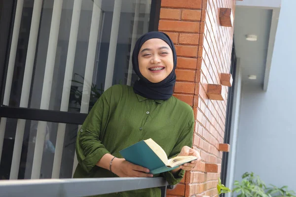 Portrait of attractive Asian hijab woman holding book in balcony. Muslim girl reading a book. Concept of literacy and knowledge