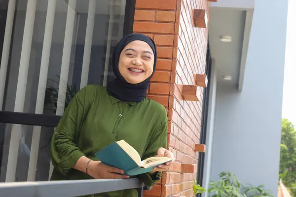 Portrait of attractive Asian hijab woman holding book in balcony. Muslim girl reading a book. Concept of literacy and knowledge