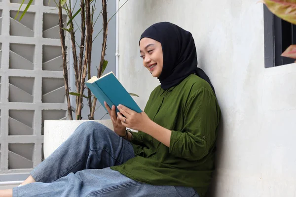 Portrait of attractive Asian hijab woman reading book in balcony. Muslim girl enjoying free time. Concept of literacy and knowledge