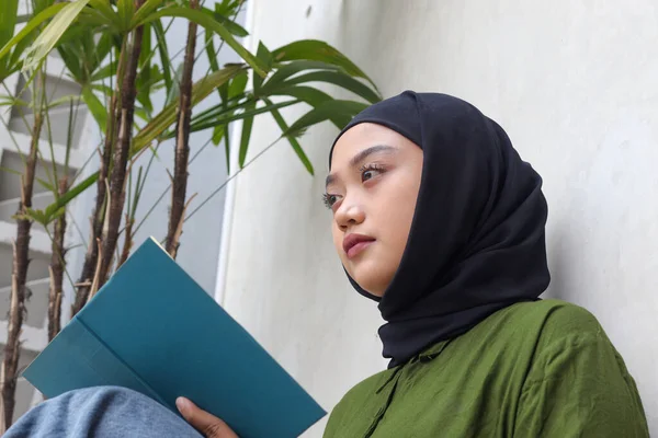 Portrait of attractive Asian hijab woman reading book in balcony. Muslim girl enjoying free time. Concept of literacy and knowledge