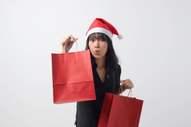 Kırmızı Noel Baba şapkalı şaşırmış Asyalı kadının portresi bir şeyler alırken mutlu oluyor. Alışveriş çantası tutan heyecanlı kız. Yeni yıl ve Noel konsepti. Beyaz arkaplanda izole edilmiş resim