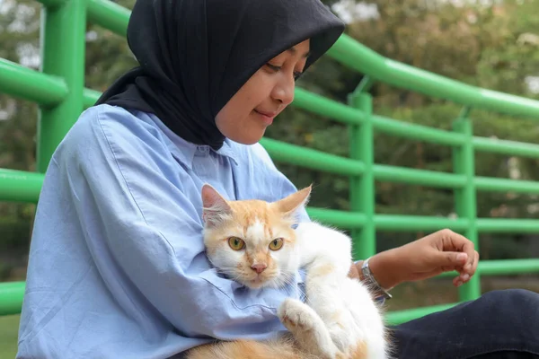 Outdoor portrait of Asian hijab woman holding and giving gentle touch to cat, taking care of her pet in nature park. Love relationship between humans and animals.