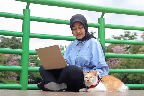Outdoor portrait of Asian hijab woman holding and giving gentle touch to cat, taking care of her pet while working on laptop in nature park. Love relationship between humans and animals.