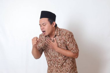 Batik tişört giyen Asyalı bir adamın portresi ve öksürürken elini avuç içinde tutan Songkok. Gri arkaplanda izole edilmiş resim