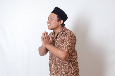 Batik tişört ve Songkok giyen gülümseyen Asyalı adamın portresi özür diliyor ve el hareketini hoş karşılıyor. Ramazan reklam konsepti. Gri arkaplanda izole edilmiş resim