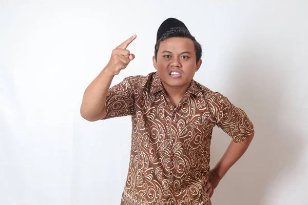 Portrait of unpleasant Asian man wearing batik shirt and songkok showing cynical unhappy angry facial expression pointing forward, giving warn. Isolated image on gray background