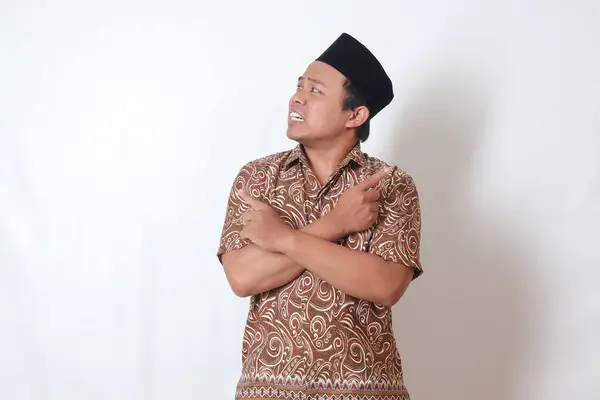 Portrait of confused Asian man wearing batik shirt and songkok with crossed hands, pointing sideways, making choice, choosing between two objects. Isolated image on gray background