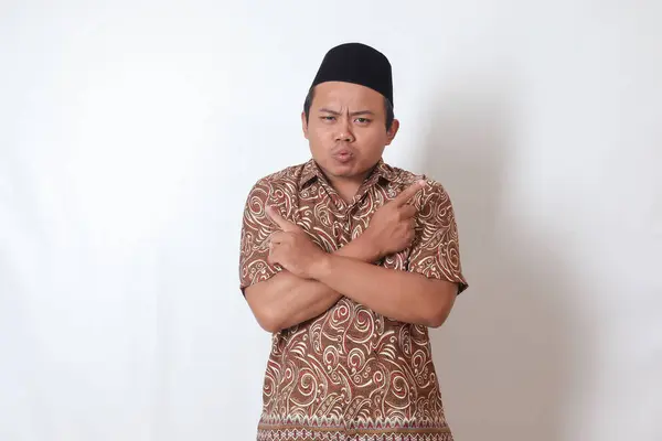 Portrait of confused Asian man wearing batik shirt and songkok with crossed hands, pointing sideways, making choice, choosing between two objects. Isolated image on gray background
