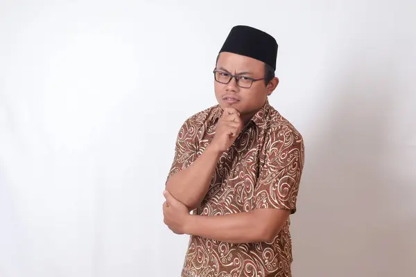Portrait of confused Asian man wearing batik shirt and songkok standing against gray background, thinking about question with hand on chin