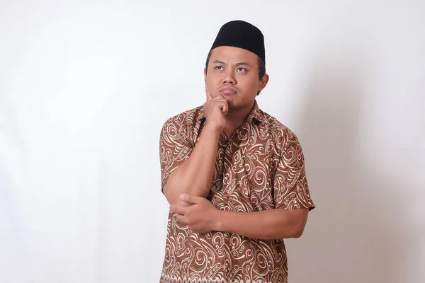 Portrait of confused Asian man wearing batik shirt and songkok standing against gray background, thinking about question with hand on chin