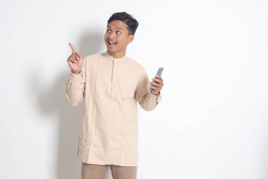Koko tişörtlü, cep telefonu tutan ve malı parmağıyla gösteren heyecanlı Asyalı Müslüman adamın portresi. Sosyal medya konsepti. Beyaz arkaplanda izole edilmiş resim