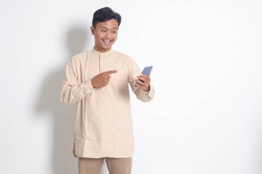 Koko tişörtlü, cep telefonu tutan ve malı parmağıyla gösteren heyecanlı Asyalı Müslüman adamın portresi. Sosyal medya konsepti. Beyaz arkaplanda izole edilmiş resim