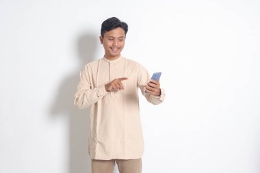 Koko tişörtlü, cep telefonu tutan ve malı parmağıyla gösteren heyecanlı Asyalı Müslüman adamın portresi. Sosyal medya konsepti. Beyaz arkaplanda izole edilmiş resim