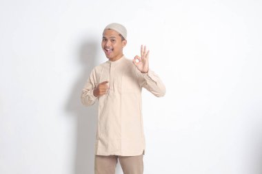 Koko tişörtlü yakışıklı Asyalı müslüman adamın portresi. Kafasında kafatası var. El hareketi yapıyor ve kameraya gülümsüyor. Onaylama konsepti. Beyaz arkaplanda izole edilmiş resim