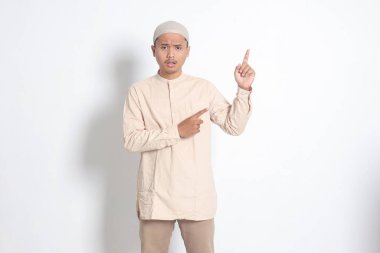 Şok olmuş Koko tişörtlü Asyalı Müslüman adamın portresi. Kafatası şapkası ile ürünü gösteriyor ve elini ve parmağını yana doğru gösteriyor. Reklam konsepti. Beyaz arkaplanda izole edilmiş resim