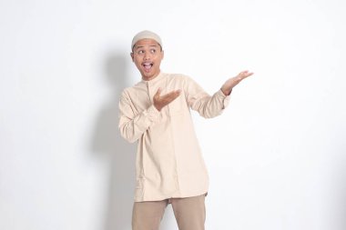 Şok olmuş Koko tişörtlü Asyalı Müslüman adamın portresi. Kafatası şapkası ile ürünü gösteriyor ve elini ve parmağını yana doğru gösteriyor. Reklam konsepti. Beyaz arkaplanda izole edilmiş resim