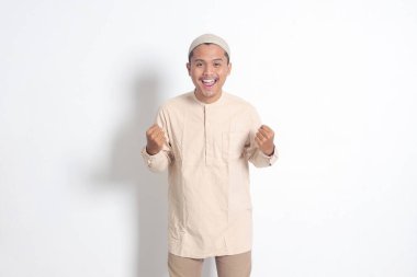 Koko tişörtlü dindar Asyalı Müslüman adamın portresi. Kafatası şapkalı. Yumruğunu kaldırıyor ve başarısını kutluyor. Başarı konsepti. Beyaz arkaplanda izole edilmiş resim