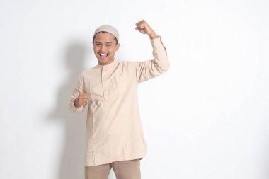 Koko tişörtlü dindar Asyalı Müslüman adamın portresi. Kafatası şapkalı. Yumruğunu kaldırıyor ve başarısını kutluyor. Başarı konsepti. Beyaz arkaplanda izole edilmiş resim