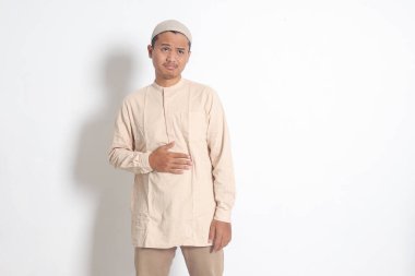 Koko tişörtlü dindar Asyalı Müslüman adamın portresi midesinde acı hisseden, açlığa dayanan. Karnım ağrıyor ve yemek konseptine açım. Beyaz arkaplanda izole edilmiş resim