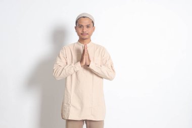 Koko tişörtlü yakışıklı Asyalı müslüman portresi. Kafatası şapkalı. Özür diliyor ve el hareketlerini hoş karşılıyor. Bayram zamanı özür dilerim. Beyaz arkaplanda izole edilmiş resim