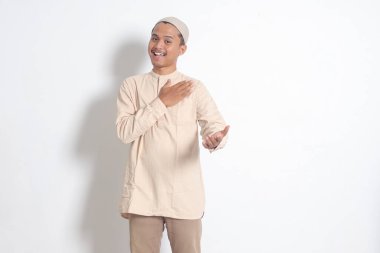 Koko tişörtlü çekici Asyalı Müslüman adamın portresi. Kafatası şapkalı ve birini içeri davet ediyor. Beyaz arkaplanda izole edilmiş resim