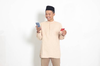 Elinde cep telefonu olan ve kredi kartı sunan Koko tişörtlü yakışıklı Asyalı Müslüman adamın portresi. Beyaz arkaplanda izole edilmiş resim