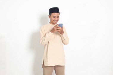 Koko tişörtlü heyecanlı Asyalı Müslüman adamın portresi. Elinde cep telefonu, yüzünde gülümseme ifadesi var. Sosyal medya ve internet kavramı. Beyaz arkaplanda izole edilmiş resim