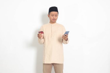 Koko tişörtlü üzgün Asyalı Müslüman adamın portresi. Elinde cep telefonu olan ve internet kredileri tarafından aldatılmış kredi kartı sunan bir kafatası. Beyaz arkaplanda izole edilmiş resim