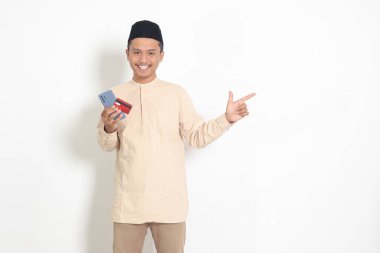 Koko tişörtlü, cep telefonu ve kredi kartı tutan Asyalı çekici Müslüman adamın portresi. Beyaz arkaplanda izole edilmiş resim