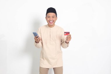Elinde cep telefonu olan ve kredi kartı sunan Koko tişörtlü yakışıklı Asyalı Müslüman adamın portresi. Beyaz arkaplanda izole edilmiş resim