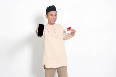 Koko tişörtlü yakışıklı Asyalı Müslüman adamın portresi. Elinde bir kredi kartı tutarken cep telefonunda model ve grafik için boş bir ekran var. Beyaz arkaplanda izole edilmiş resim