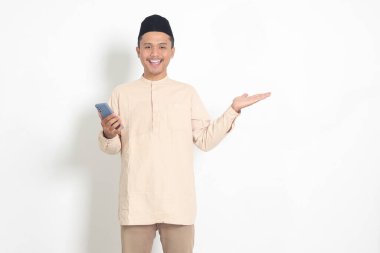 Koko tişörtlü yakışıklı Asyalı Müslüman adamın portresi. Kafasında cep telefonu olan, malı parmağıyla gösteren ve gösteren. Sosyal medya konsepti. Beyaz arkaplanda izole edilmiş resim