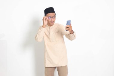 Kafasında cep telefonu olan Koko gömlekli, şaşırmış Asyalı Müslüman adamın portresi. Şok olmuş yüz ifadesi sergiliyor. Reklamcılık ve sosyal medya kavramı. Beyaz arkaplanda izole edilmiş resim