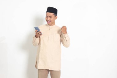 Koko tişörtlü heyecanlı Asyalı Müslüman adamın portresi. Kafatası şapkalı, yumruğunu kaldırıyor ve maçı cep telefonuyla kazanıyor. Reklamcılık ve sosyal medya kavramı. Beyaz arkaplanda izole edilmiş resim