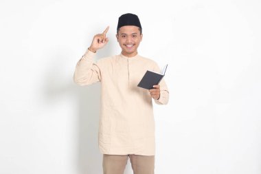 Koko tişörtlü çekici Asyalı Müslüman adamın portresi. Peci kitabı okuyor. Parmakla ve kalemle işaret ederken bir fikri olduğunu söylüyor. Beyaz arkaplanda izole edilmiş resim