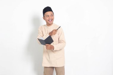 Koko tişörtlü heyecanlı müslüman adamın portresi. Peci bir defter okuyor. Mutlu Asyalı adam parmağıyla kenarı işaret ediyor. Beyaz arkaplanda izole edilmiş resim
