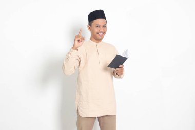 Koko tişörtlü çekici Asyalı Müslüman adamın portresi. Peci kitabı okuyor. Parmakla ve kalemle işaret ederken bir fikri olduğunu söylüyor. Beyaz arkaplanda izole edilmiş resim