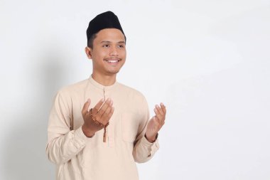 İslami boncuklar tutan, elleri havada dua eden, Koko tişörtlü, dindar Asyalı Müslüman adamın portresi. Dindar inanç konsepti. Beyaz arkaplanda izole edilmiş resim