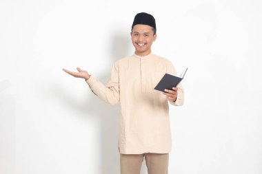 Koko tişörtlü heyecanlı müslüman adamın portresi. Peci bir defter okuyor. Mutlu Asyalı adam parmağıyla kenarı işaret ediyor. Beyaz arkaplanda izole edilmiş resim
