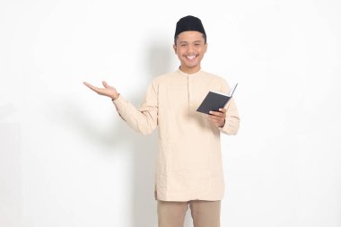 Koko tişörtlü heyecanlı müslüman adamın portresi. Peci bir defter okuyor. Mutlu Asyalı adam parmağıyla kenarı işaret ediyor. Beyaz arkaplanda izole edilmiş resim