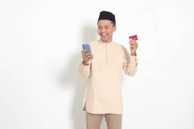 Elinde cep telefonu olan ve kredi kartı sunan Koko tişörtlü yakışıklı Asyalı Müslüman adamın portresi. Beyaz arkaplanda izole edilmiş resim