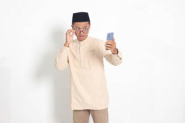 Kafasında cep telefonu olan Koko gömlekli, şaşırmış Asyalı Müslüman adamın portresi. Şok olmuş yüz ifadesi sergiliyor. Reklamcılık ve sosyal medya kavramı. Beyaz arkaplanda izole edilmiş resim