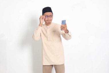 Kafasında cep telefonu olan Koko gömlekli, şaşırmış Asyalı Müslüman adamın portresi. Şok olmuş yüz ifadesi sergiliyor. Reklamcılık ve sosyal medya kavramı. Beyaz arkaplanda izole edilmiş resim