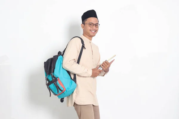 Portrait of excited student Asian muslim man in koko shirt with skullcap carrying backpack, while holding his school books. Islamic education concept. Isolated image on white background