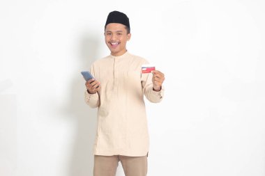 Elinde cep telefonu olan ve kredi kartı sunan Koko tişörtlü yakışıklı Asyalı Müslüman adamın portresi. Beyaz arkaplanda izole edilmiş resim