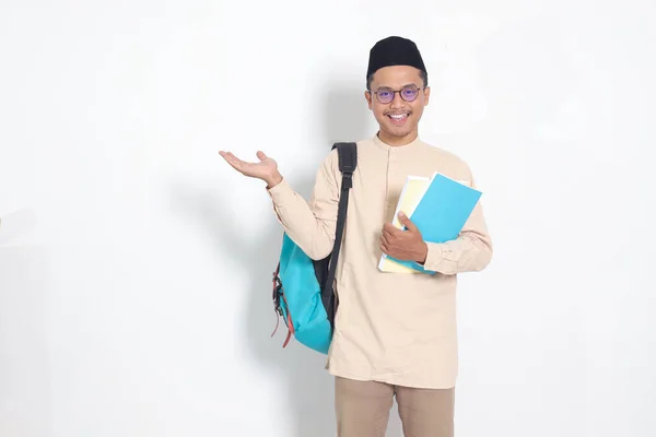 Portrait of excited student Asian muslim man in koko shirt with skullcap carrying backpack, holding school books, pointing to the side. Islamic education concept. Isolated image on white background