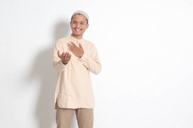 Koko tişörtlü çekici Asyalı Müslüman adamın portresi. Kafatası şapkalı ve birini içeri davet ediyor. Beyaz arkaplanda izole edilmiş resim