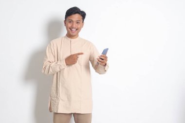 Koko tişörtlü, cep telefonu tutan ve malı parmağıyla gösteren heyecanlı Asyalı Müslüman adamın portresi. Sosyal medya konsepti. Beyaz arkaplanda izole edilmiş resim
