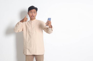 Koko tişörtlü heyecanlı Asyalı Müslüman adamın portresi. Elinde cep telefonu, fotoğrafını çekiyor, selam veriyor ve elini sallıyor. Sosyal medya konsepti. Beyaz arkaplanda izole